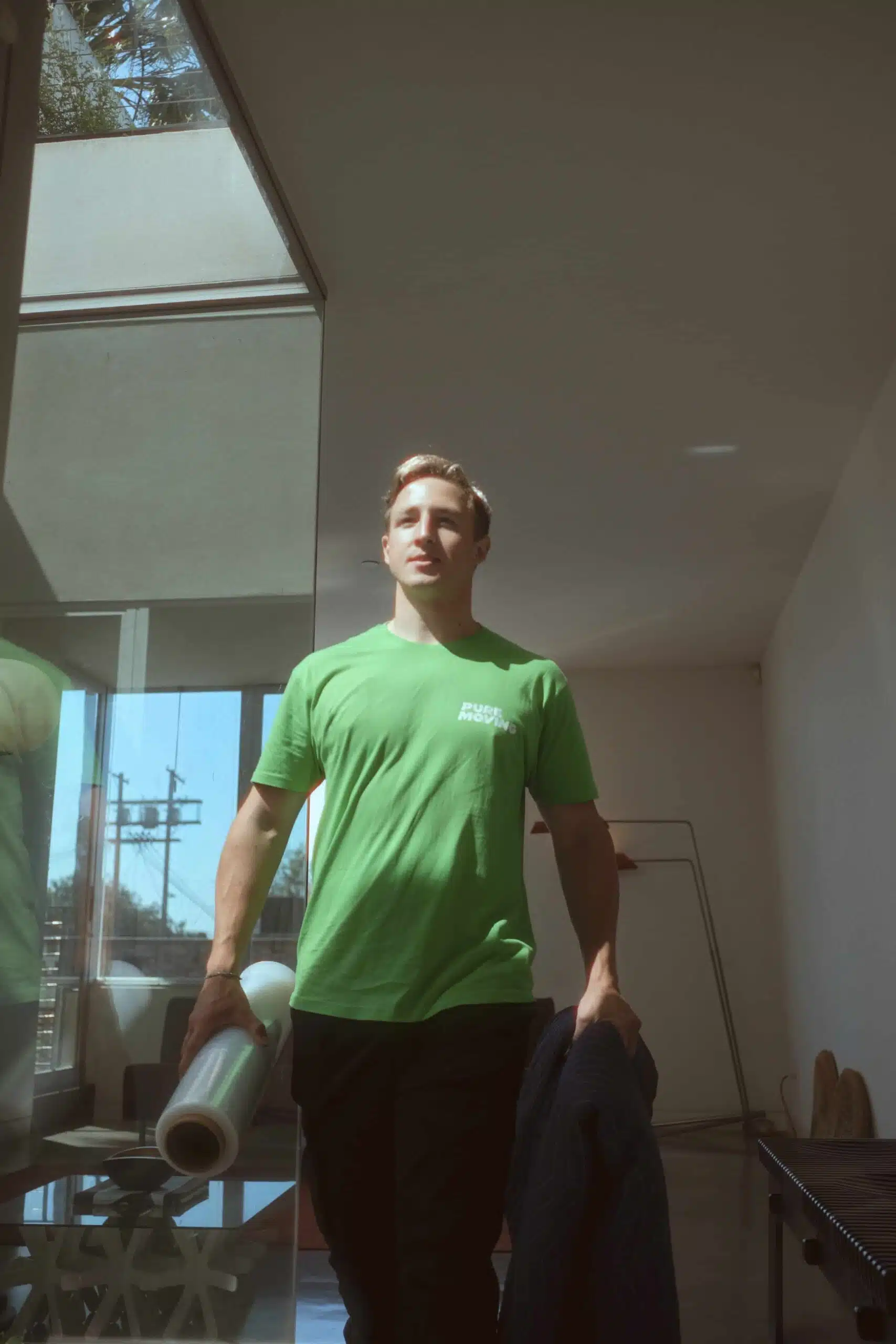 A person wearing a green shirt and holding a rolled-up piece of paper and a jacket walks through a well-lit room with large windows, resembling an efficient commercial moving service office.