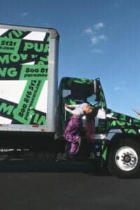 A person with long hair and colorful attire is hanging onto the door of a green and white moving truck with 