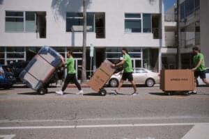 Three movers in green shirts, offering top-notch Moving and Packing Services, push items on dollies across a street in front of a modern apartment building.