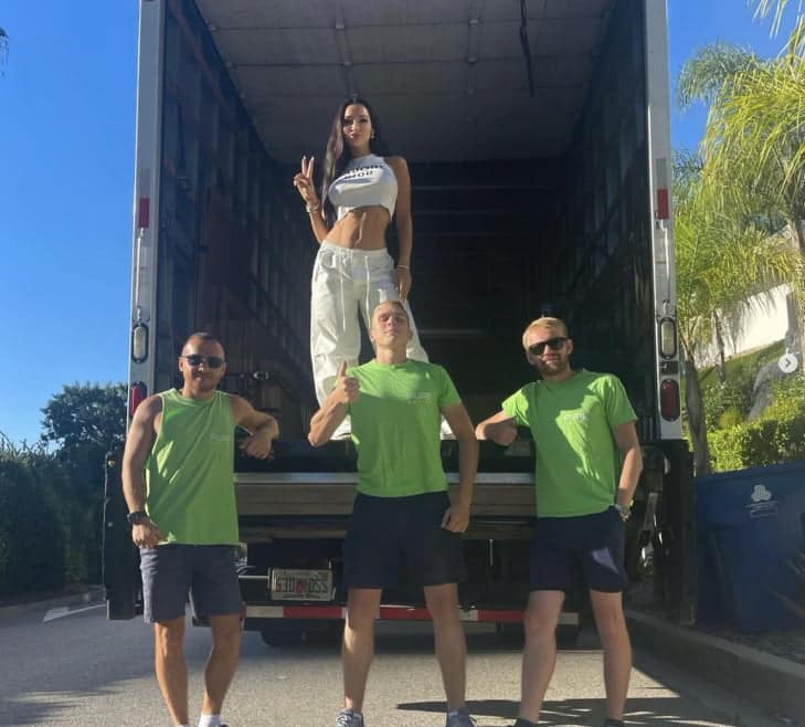 Three men in green shirts and shorts stand in front of a truck, with a woman in white clothing posing inside the truck. The woman, perhaps moving from Los Angeles to Miami, is making a peace sign.