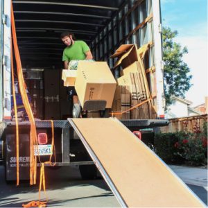 A man in a green shirt uses a hand truck to move boxes into the back of a moving truck parked on a residential street, with a ramp extending down to the ground as he prepares for the long journey moving from Los Angeles to Miami.