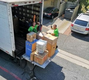Two movers in green shirts stand on the back of a truck filled with boxes, efficiently organizing items for a Full Service Moving operation on a residential street.