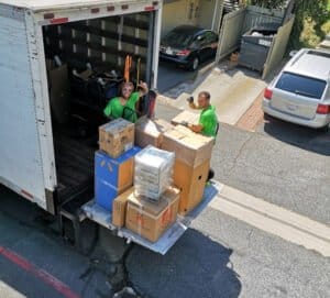 Two movers in green shirts load boxes onto a moving truck parked in a residential street. The truck's back ramp is open with several packed boxes on it.