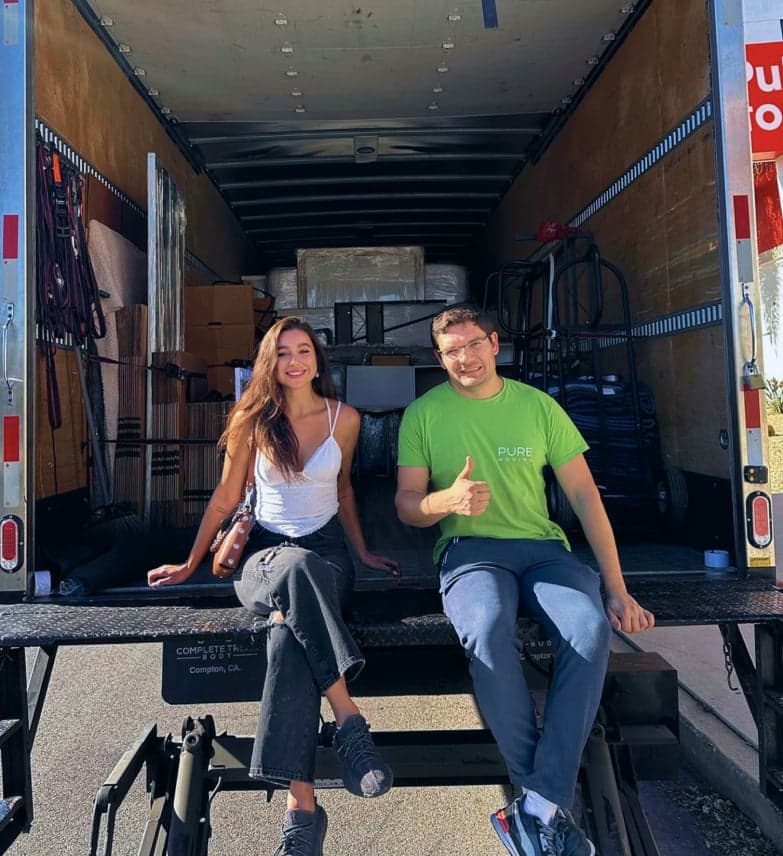Two people are sitting on the tailgate of a moving truck filled with boxes and furniture. The woman is smiling while the man, perhaps thinking about Bay Area movers, gives a thumbs up.