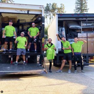 Six men in green shirts stand on and around the back of two moving trucks loaded with items. One man holds a green t-shirt that reads 