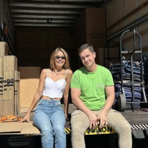 Two people sitting on the edge of a moving truck, one wearing a white top and jeans, the other in a green shirt and khaki pants, with moving supplies and information about moving rates in the background.