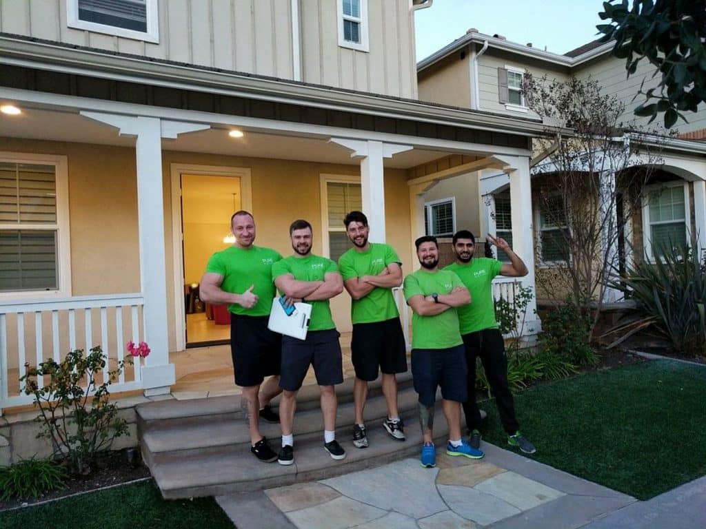 Five Pure movers in green t-shirts pose confidently in front of a house, each folding their arms, with one holding a clipboard.