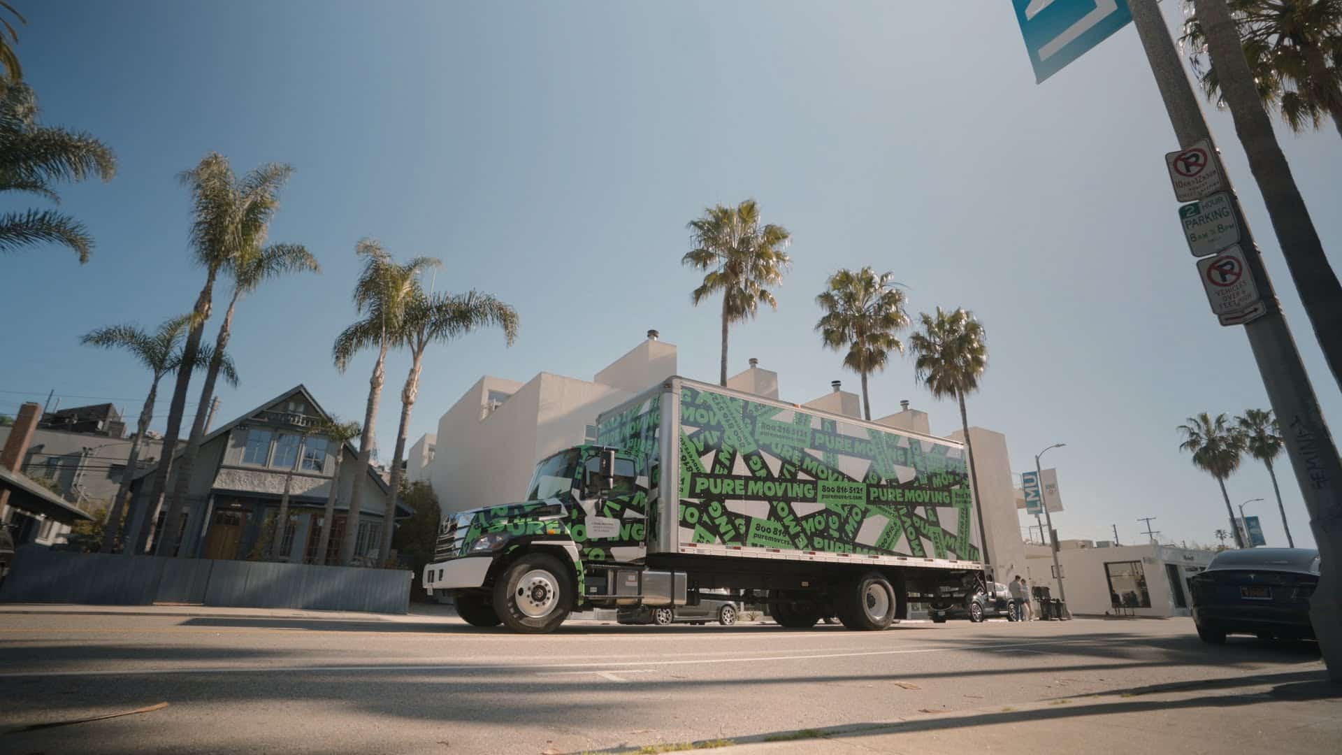 A sunny street scene with tall palm trees and a moving company truck turning at an intersection.