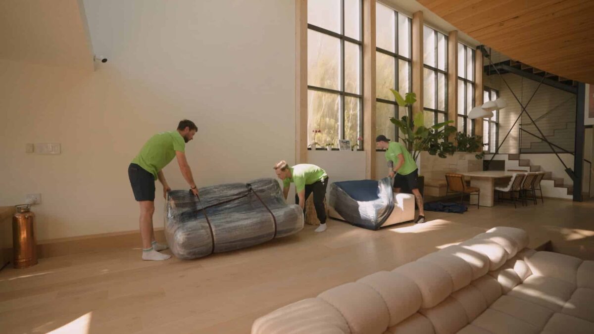 Three people in green shirts are moving wrapped furniture in a bright living room with large windows and a leather couch, carefully following their packing checklist. Long distance movers
