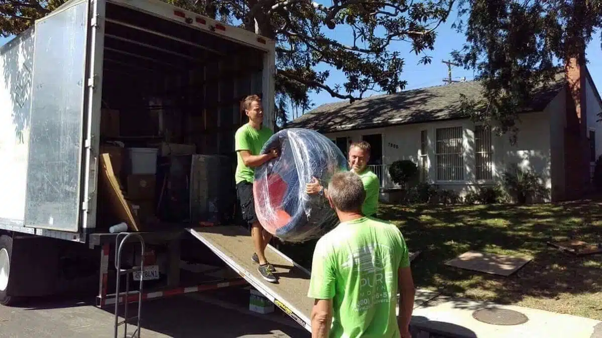 Three movers from a moving company unloading a wrapped mattress from a truck in front of a house on a sunny day. Long Beach